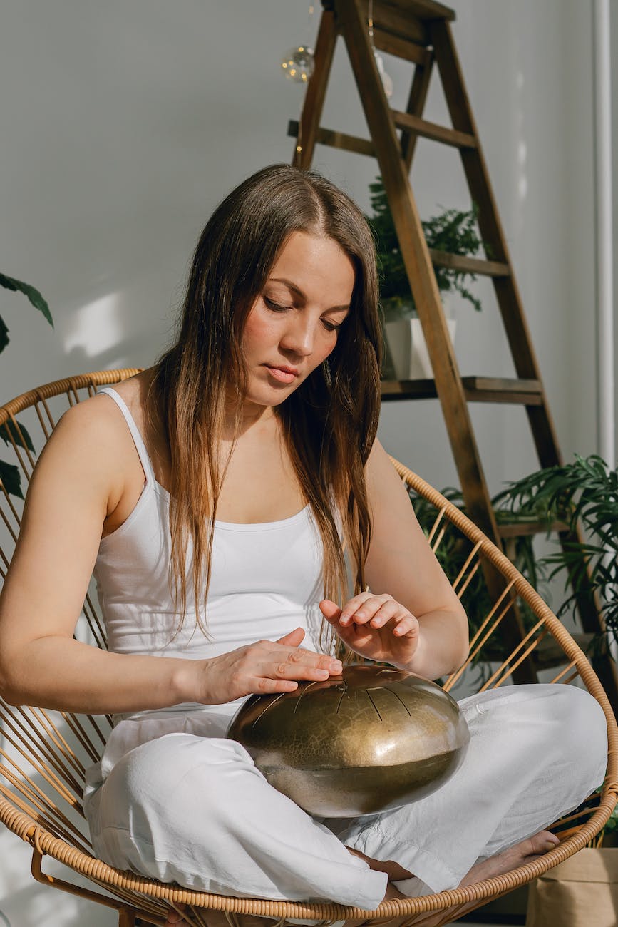 a woman in white tank top sitting on a woven chair while playing a steel tongue drum