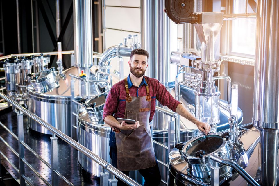 Man with a brown leather apron holding a tablet standing next to a large beer fermentation tank inside a brewery.