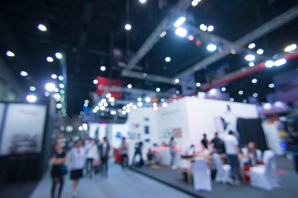 A trade show floor with several trade show booths set up and many visitors walking around each booth. The image is slightly out of focus, obscuring some details.