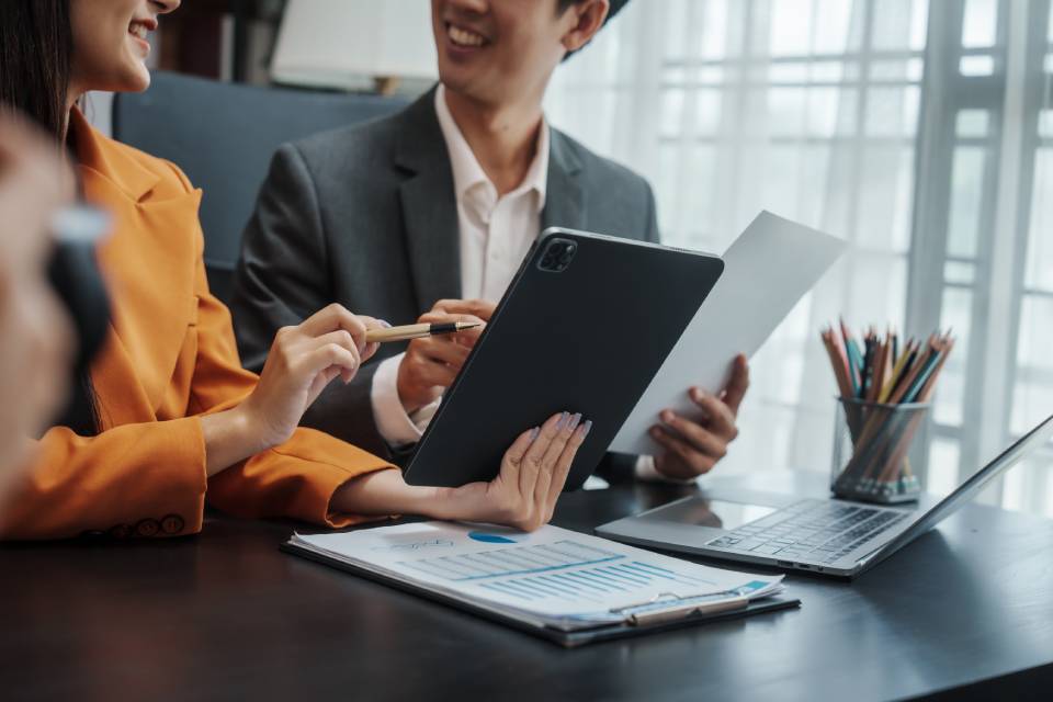 Two businesspeople are sitting at a desk with tablets and a laptop while discussing information on a spreadsheet.