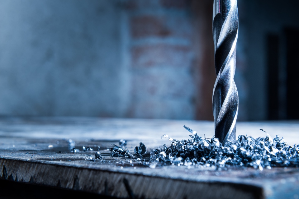 A drill and metal shavings, up close, against a concrete and brick backdrop. The drill hangs above the metal.