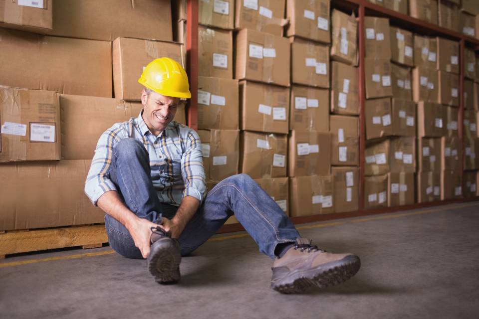 A warehouse worker with a pained expression is on the floor, leaning against a full pallet rack. He clutches his ankle, which is sprained.