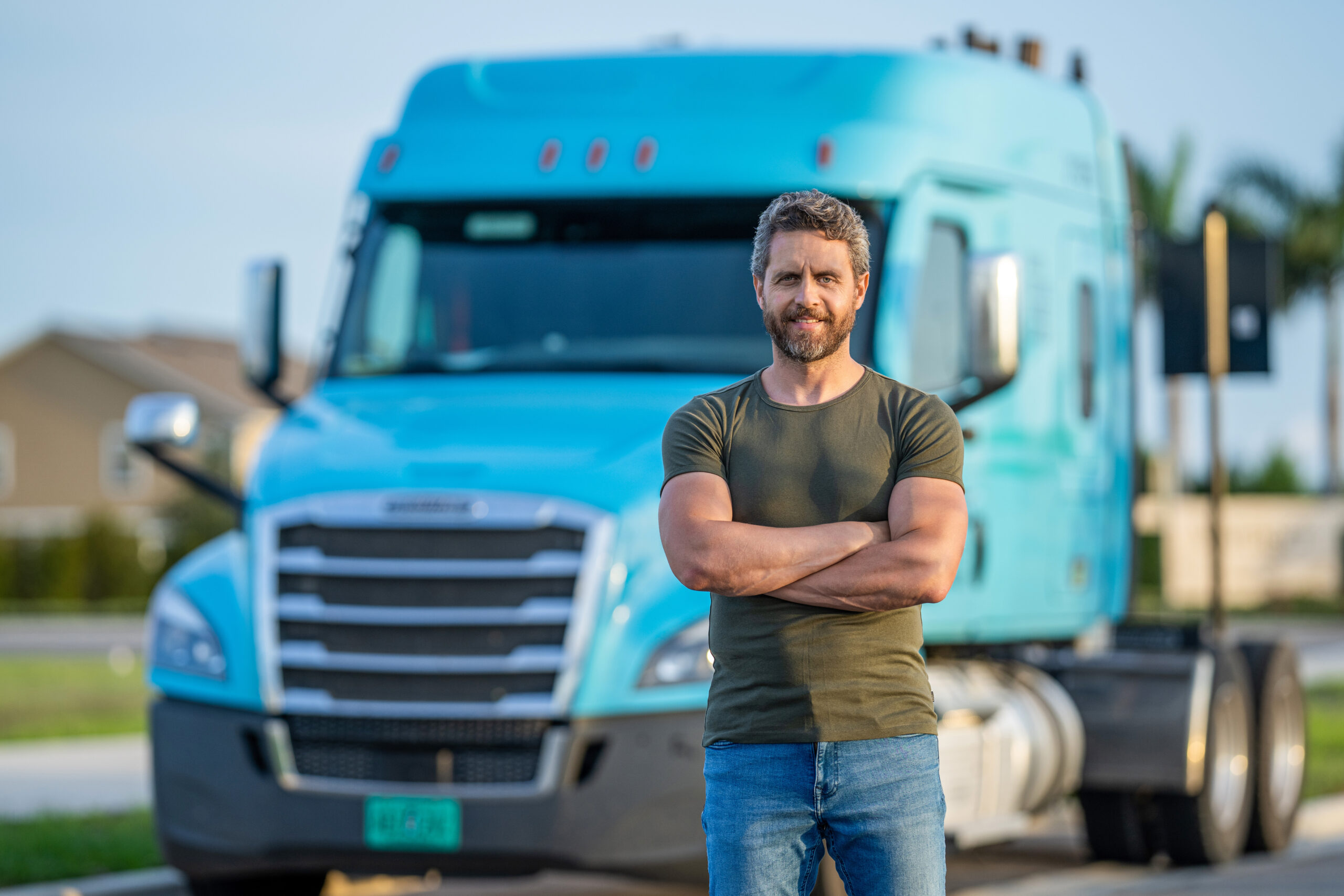 A man wearing an olive green shirt and jeans stands in front of a turquoise semi-truck cab. He has brown hair.