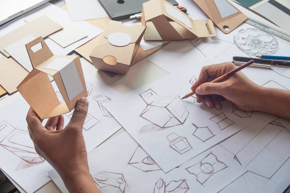 Closeup view of a designer sketching out brand product packaging while holding a prototype of a cardboard box.