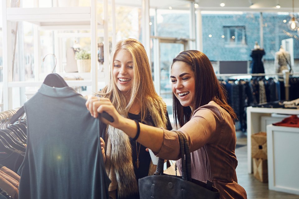 Two young women are shopping at a clothing boutique. They are holding up a shirt on a hanger and admiring it.