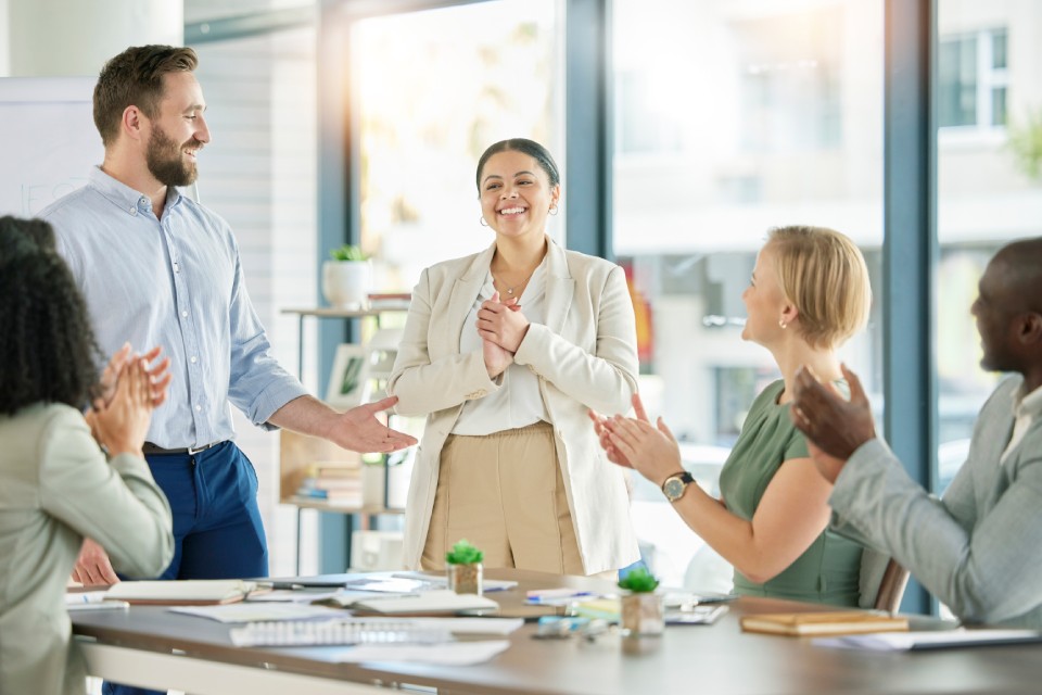 A smiling woman accepts praise and applause from her manager and colleagues who sit around a table near her.