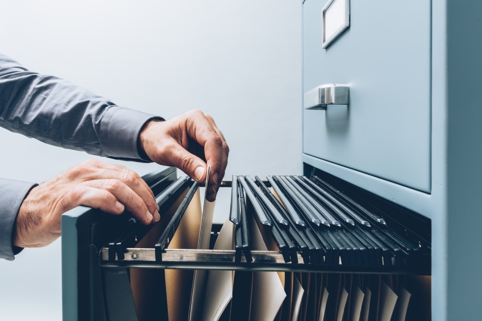 Closeup side view of a man’s hands searching for a file in a metal filing cabinet with hanging file folders.