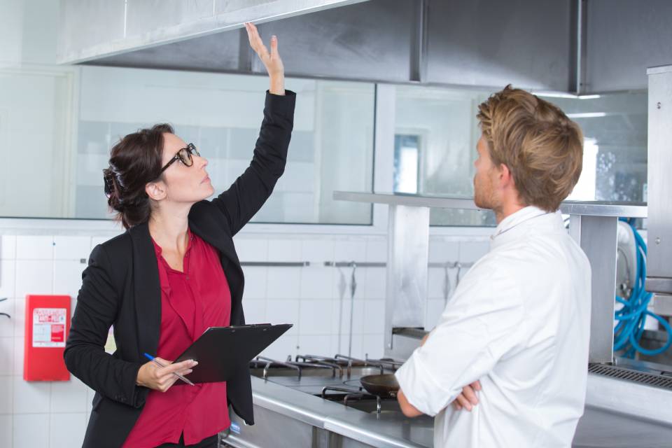 A woman with a clipboard inspecting the ventilation hood in a restaurant kitchen while the chef looks on.