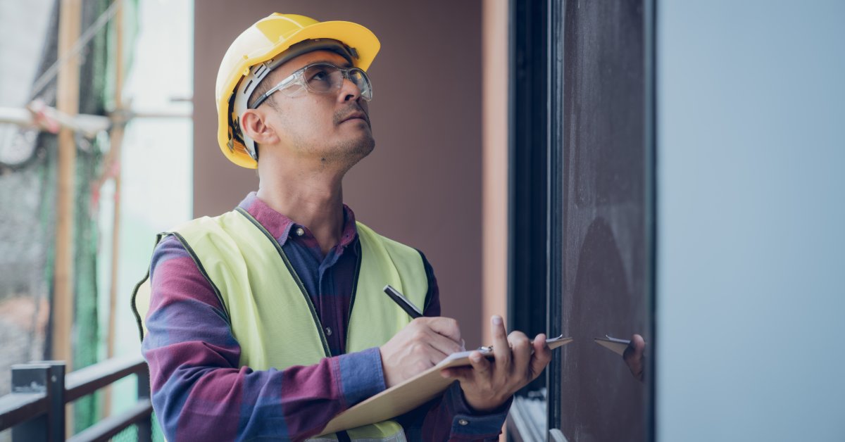 A man wearing a hard hat, safety glasses, and a yellow vest conducts a safety inspection with a pen and clipboard.