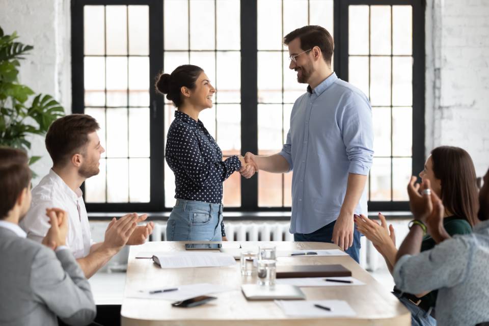 A group of people clapping around a conference table. Up front, a man and woman stand up to shake hands, grinning.