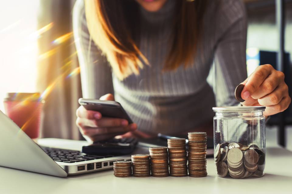 Close-up of a woman holding a smartphone with her right hand and putting coins inside a jar with her left hand over a table.