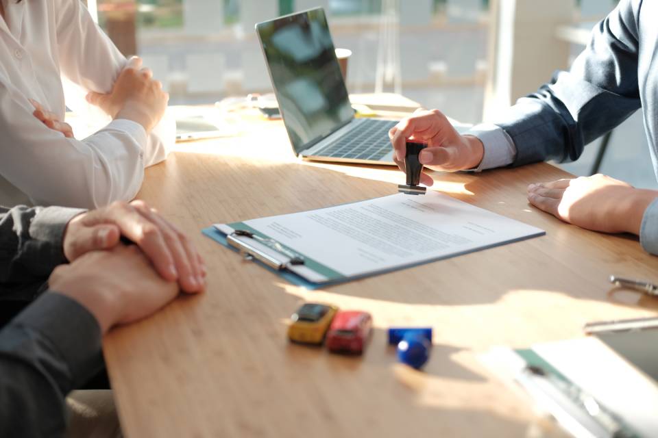 Three people sitting at a desk with a laptop and clipboard. A man in a suit is stamping a form on the clipboard.