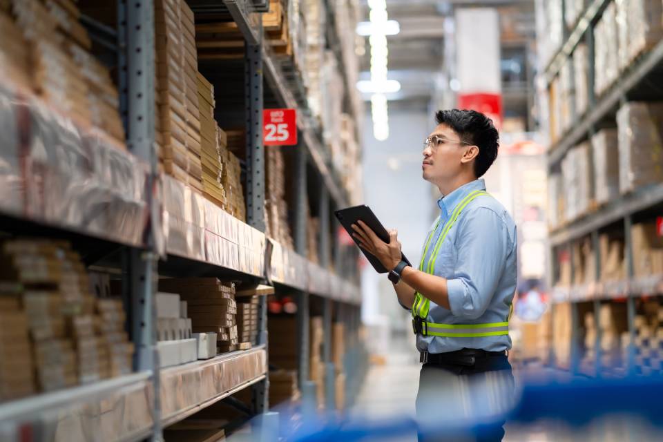 A man wearing a blue shirt and a yellow safety belt. He holds a tablet and examines shelves of inventory in a warehouse.