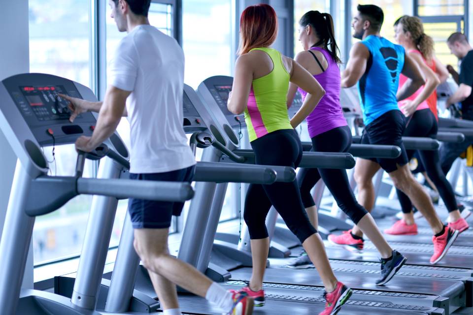 A group of three men and three women are running on different treadmills that face large windows at a gym.