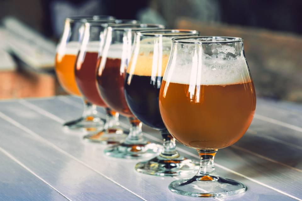 Five large, round glasses filled with beers of different shades in a straight line on top of a metal surface.