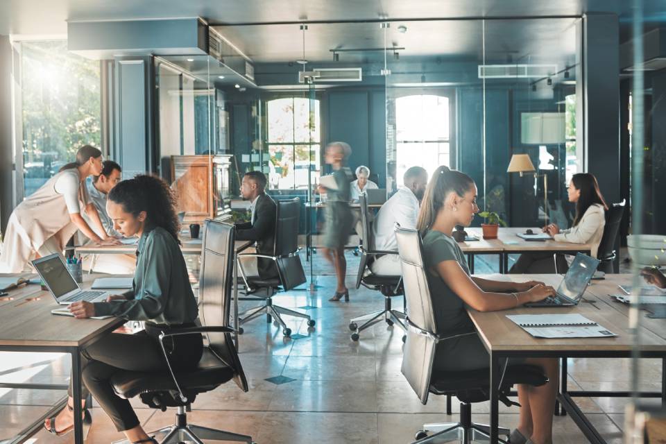 Employees sit in desk chairs at wooden tables and work on laptops in a busy open-floor plan office space.