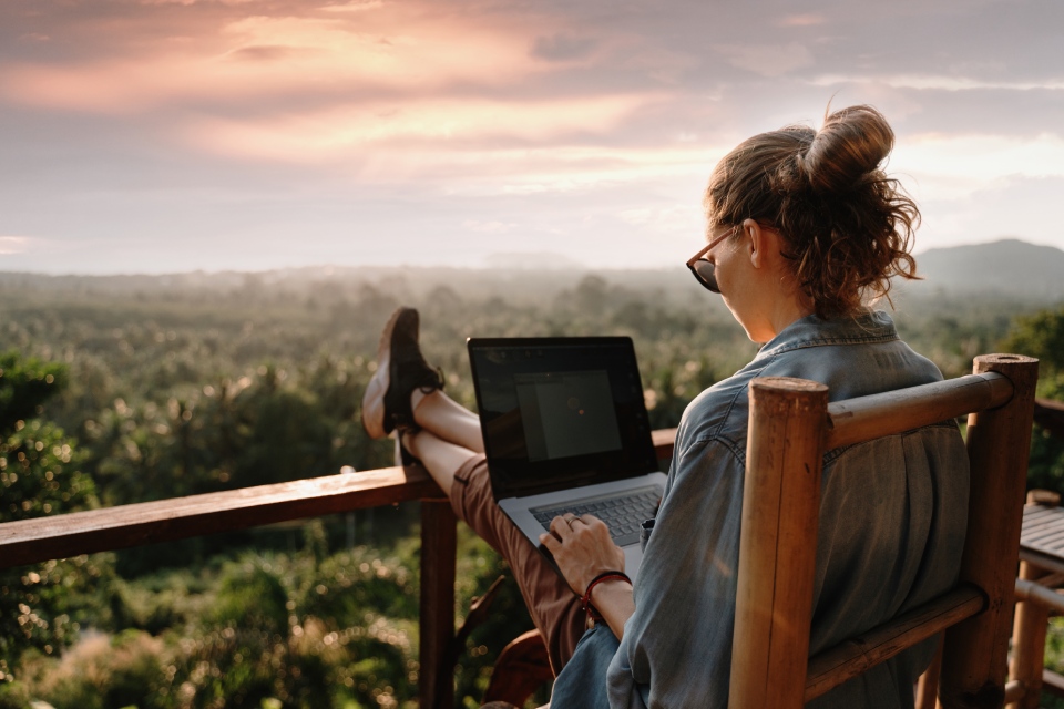 A woman is sitting outside on a deck with her feet up as she works on a silver laptop while the sun sets over the trees.