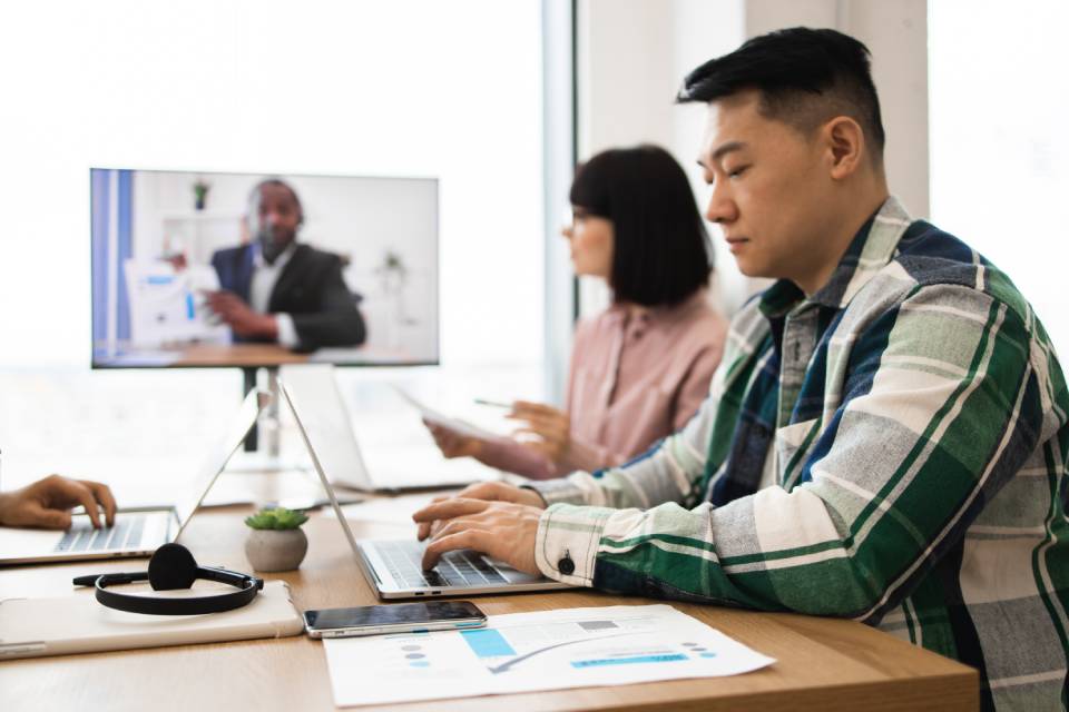 Two employees in business-casual clothing sit at a table in front of laptops. Someone on a screen is talking in the background.