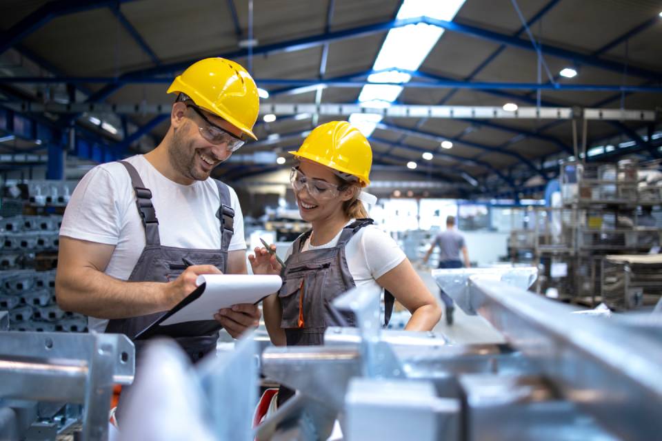 Two factory workers looking over paperwork on a clipboard. They are both wearing hard hats and overalls.