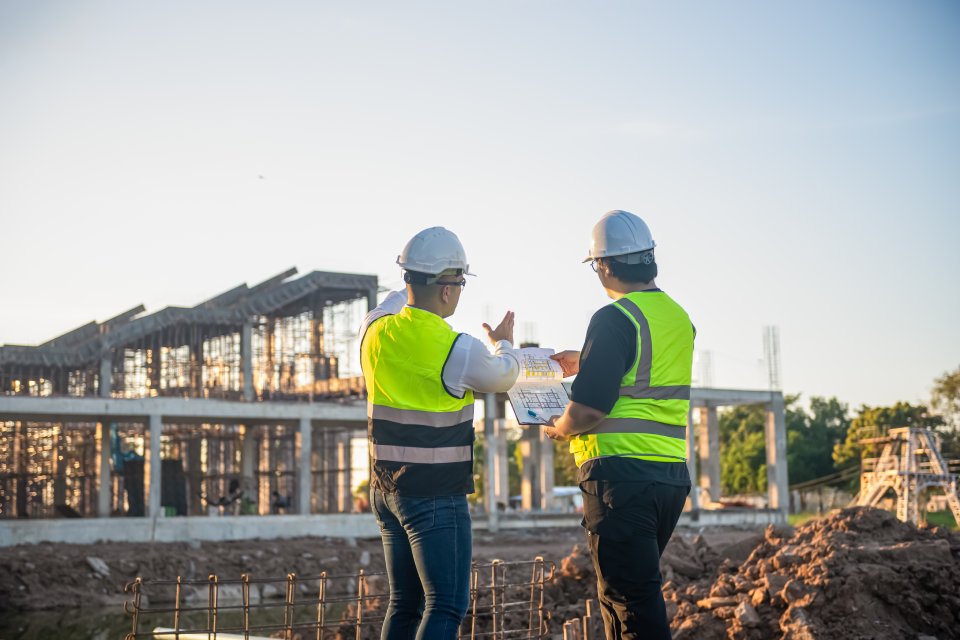 Two men in safety vests and white hard hats stand near a construction project while looking at a clipboard.