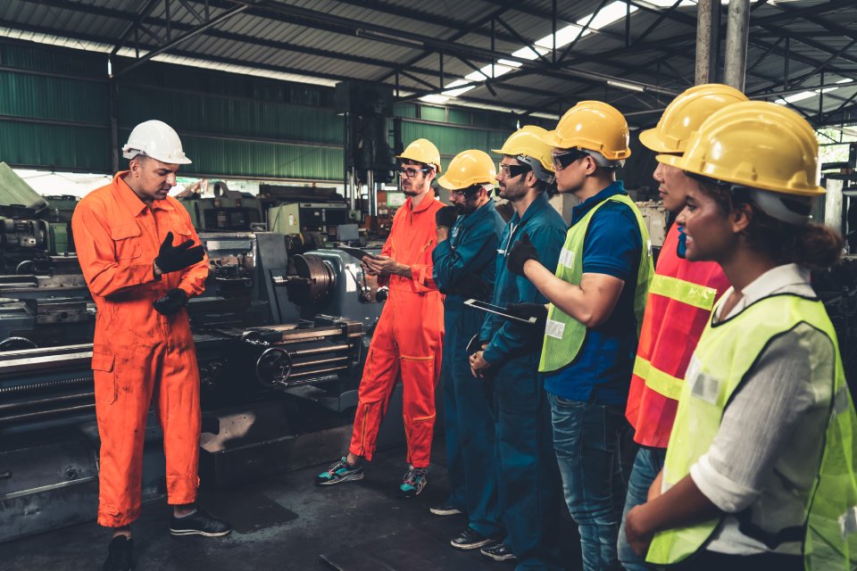 A group of seven workers wearing protective helmets and high-visibility gear stand together in an industrial area.