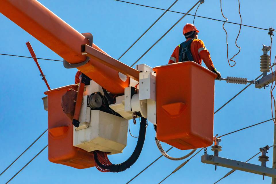 A worker operates an orange bucket lift while looking at overhead power lines against a clear blue sky.