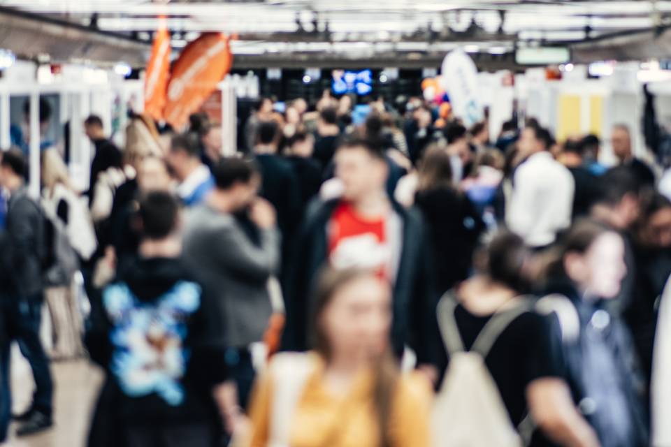 A blurred image of a crowd of people walking around in a convention center at a business trade show.