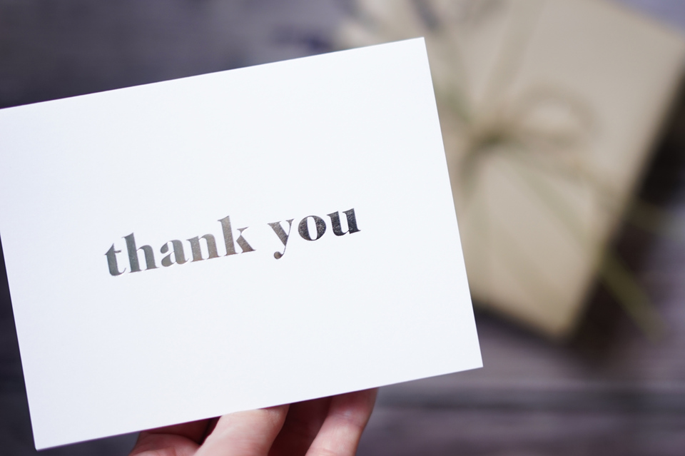 A person's hand holding a white card reading "thank you" in black text. A wrapped present sits on a wooden background.