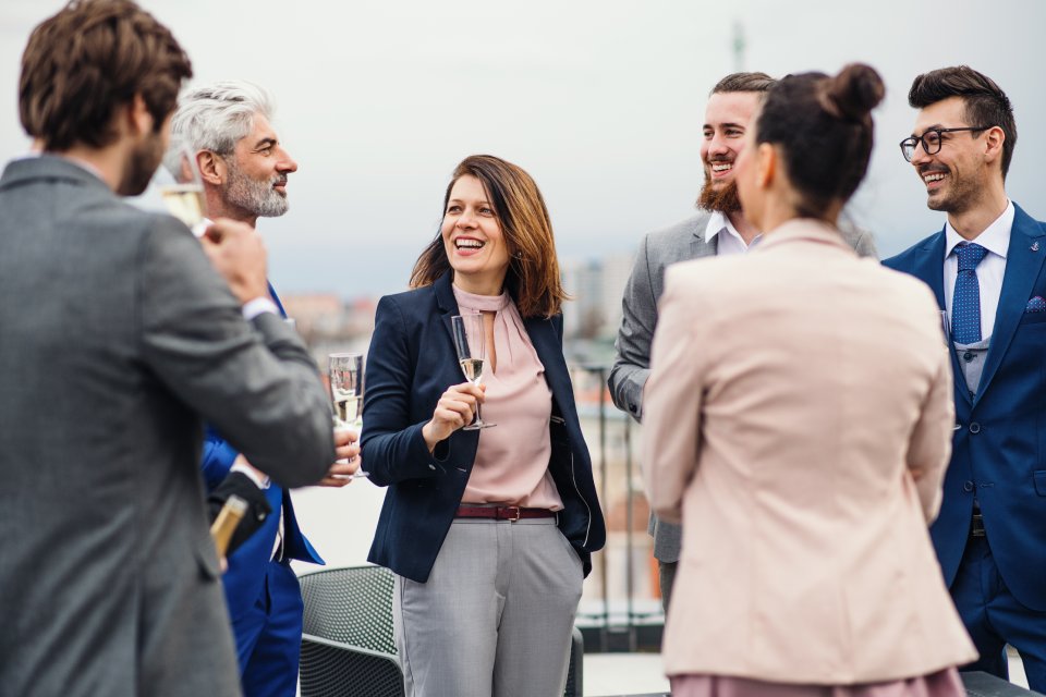 A group of smiling people stand on an outdoor terrace. They're wearing professional clothing and drinking champagne.