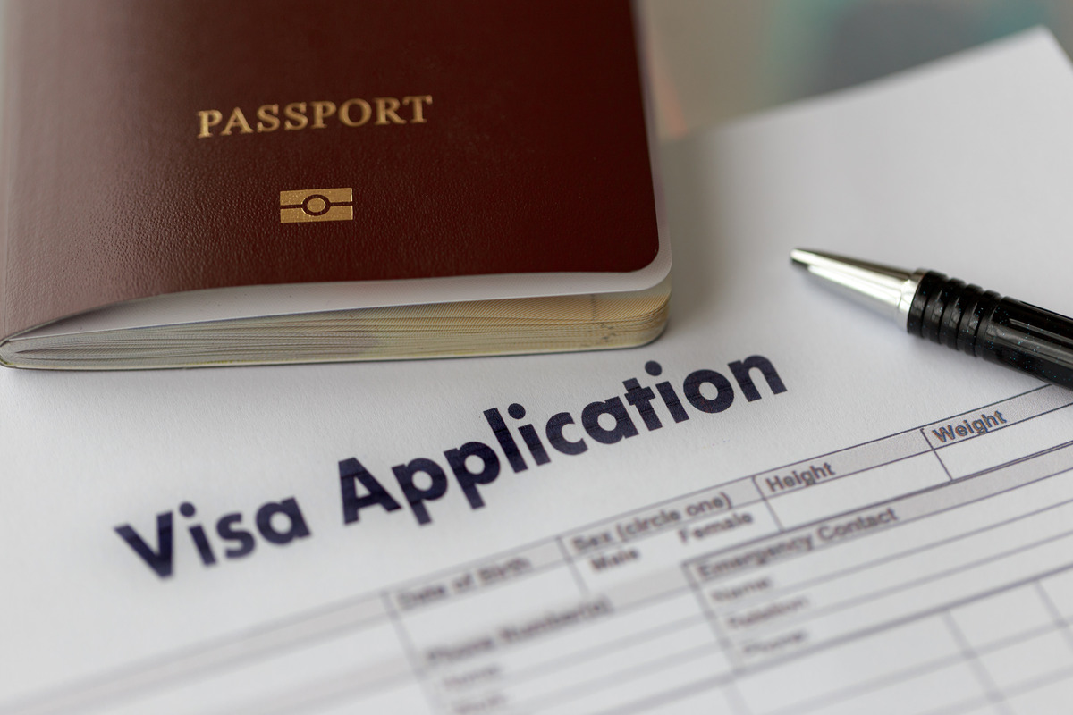 A brown passport and a black pen are resting on top of a piece of paper that says "Visa Application" in black text.