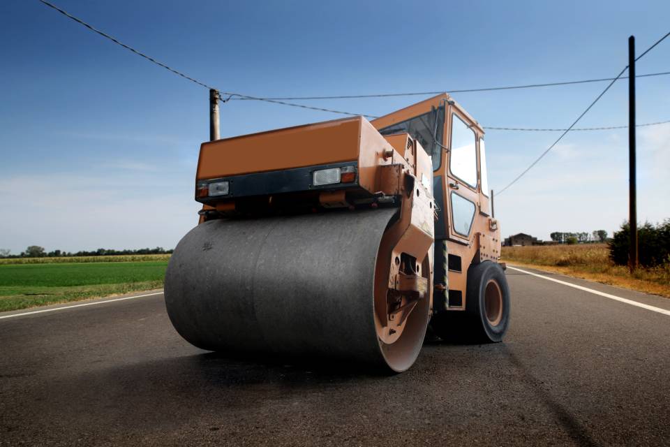 An orange asphalt paving machine with large rollers sits parked diagonally on black asphalt under a clear blue sky.