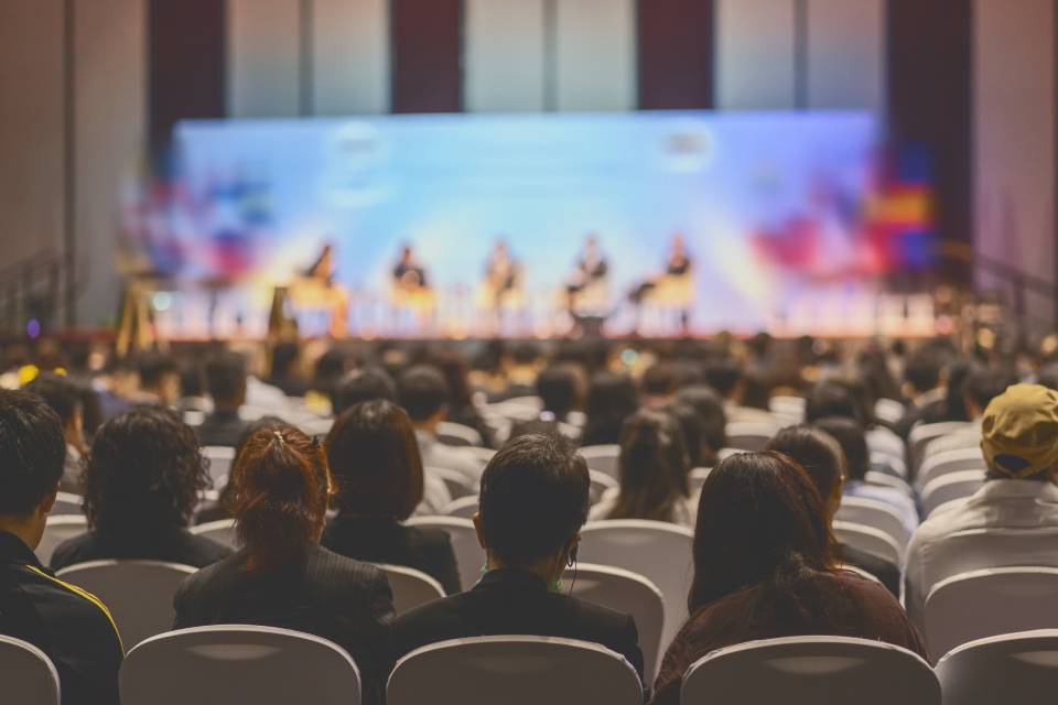 A group of speakers sit on a stage in the background with a large crowd watching in rows of chairs from the audience.