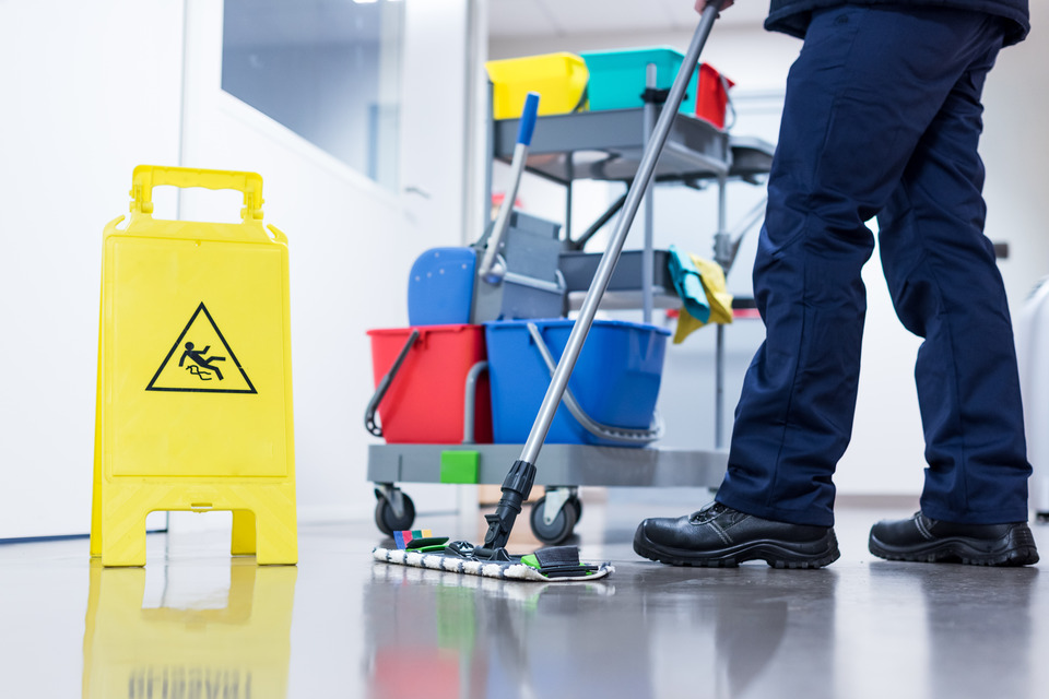 An employee in blue pants pushes a dry mop across the floor next to a cleaning cart and yellow wet floor sign.