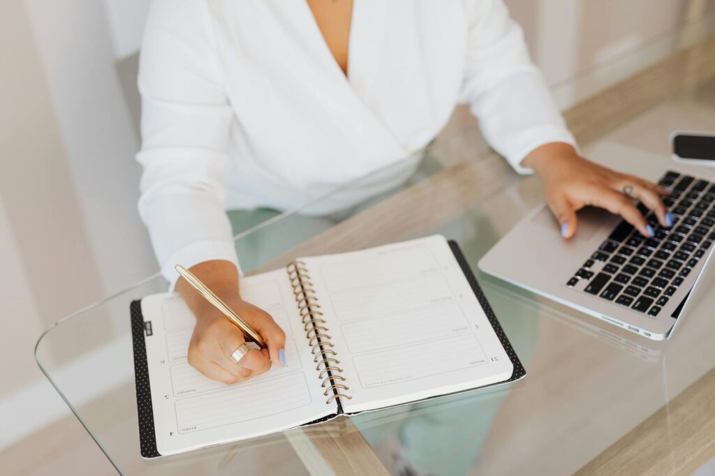person writing on notebook while using a laptop
