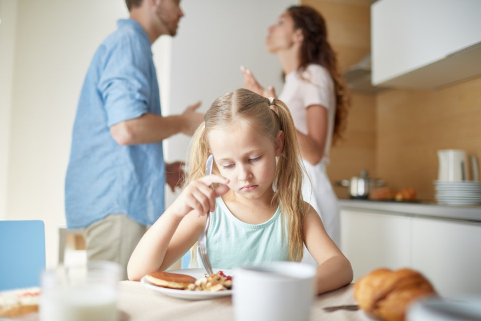 An sad girl trying to eat her breakfast at the kitchen table while her blurred parents argue behind her.