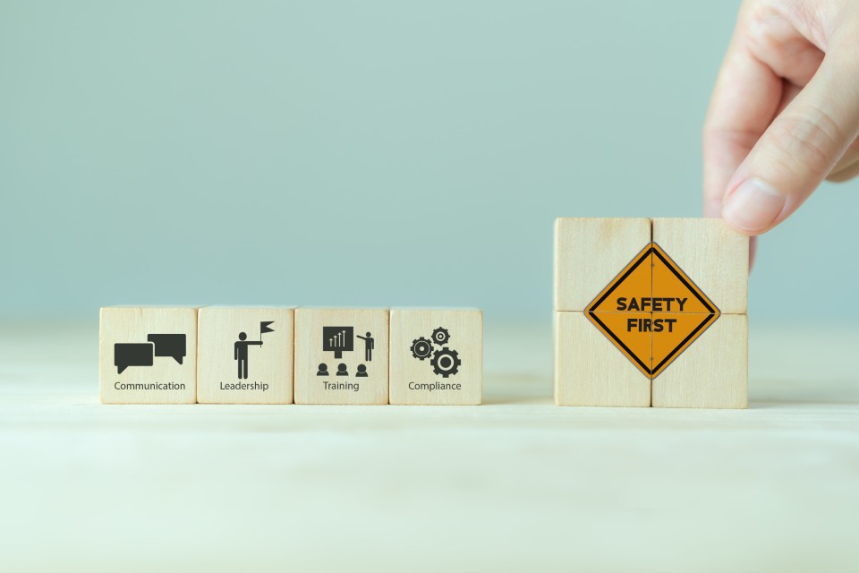 A hand touches small wooden blocks that form the words "SAFETY FIRST" next to four other blocks with business terms.
