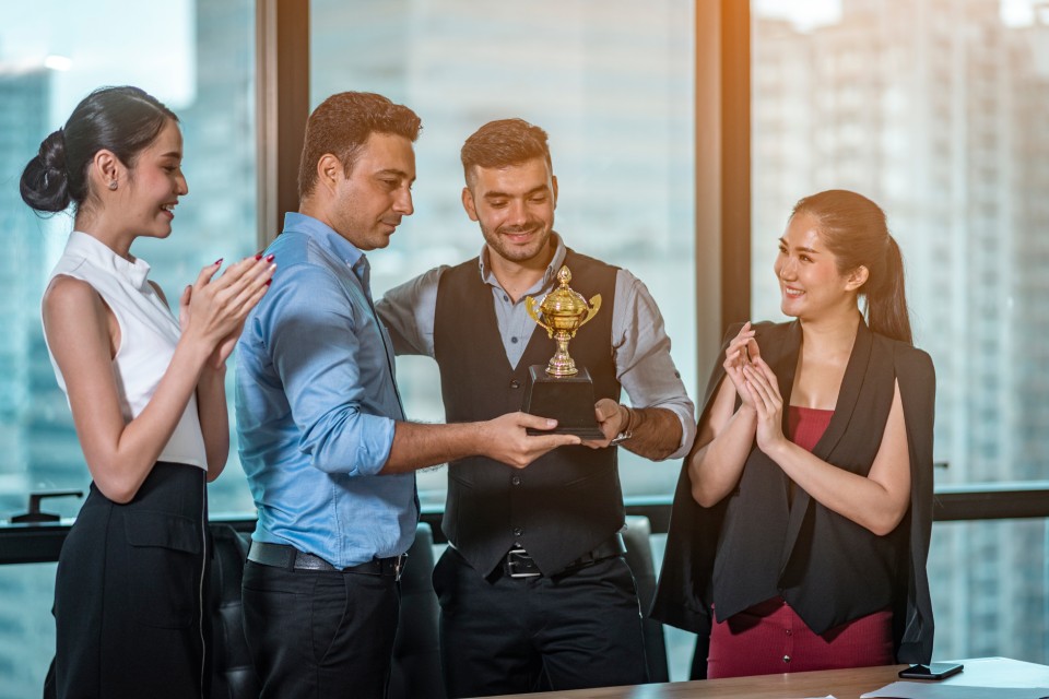 A group of four people stand together in front of a large window as they look at a small golden trophy.
