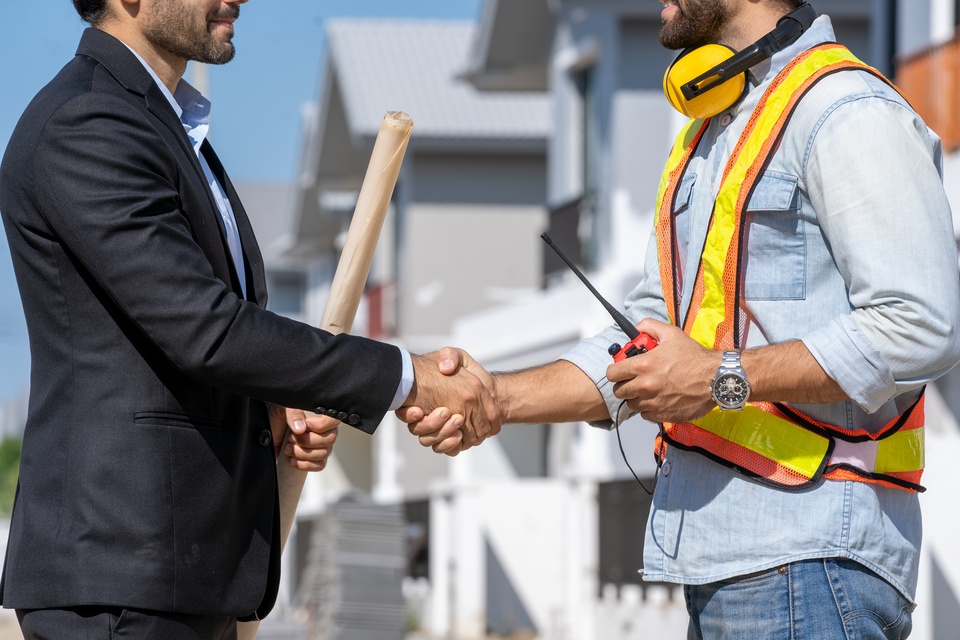 An engineer wearing a high-visibility vest and a radio shaking hands with a man in a suit on a construction site.