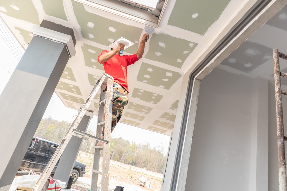 A low-level shot of an owner on a ladder using taping knives to seal drywall sheets on the ceiling.