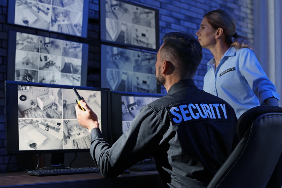 Two security guards sitting at a desk wearing shirts that say "SECURITY". They're watching a wall of camera monitors.