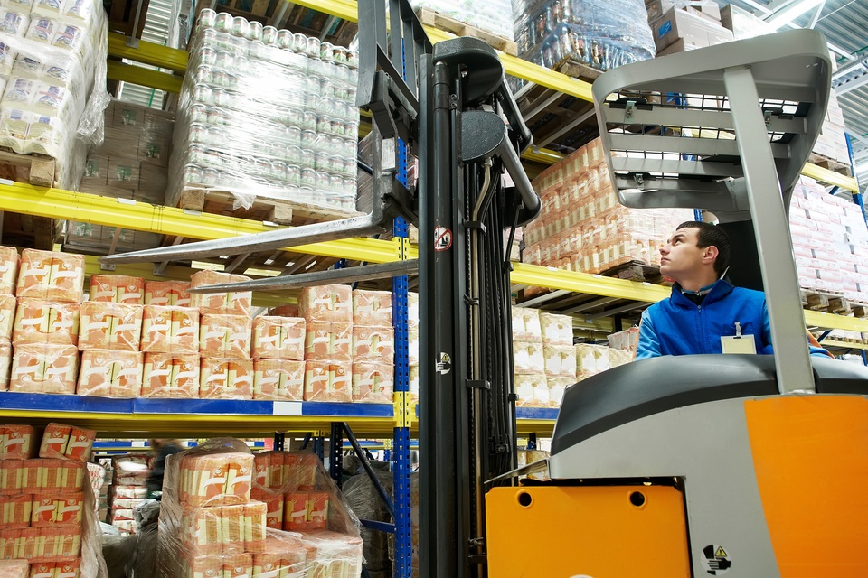 A warehouse worker operating a forklift while looking up at a pallet of products sitting on a shelf.