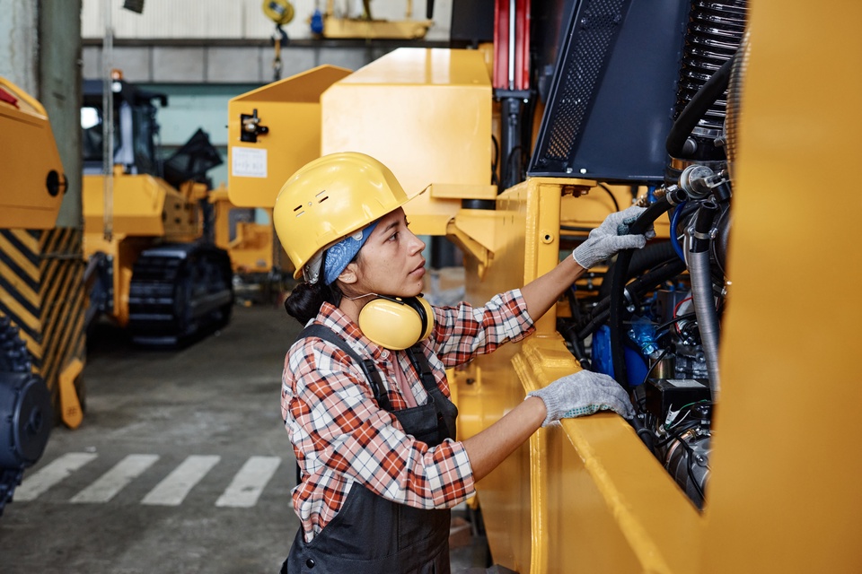 A young woman wearing a hard hat and gloves is examining the side control panel of a large piece of construction equipment.