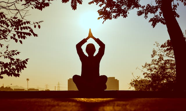 yoga pose-meditation A person practicing yoga outdoors at sunrise, with hands in a prayer position, as they focus on their breath to secure stability and inner peace