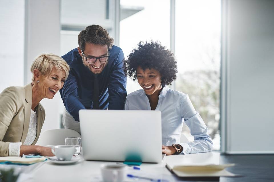 Two smiling women and a smiling man gather around a laptop at work. The man is standing, and the women are sitting.