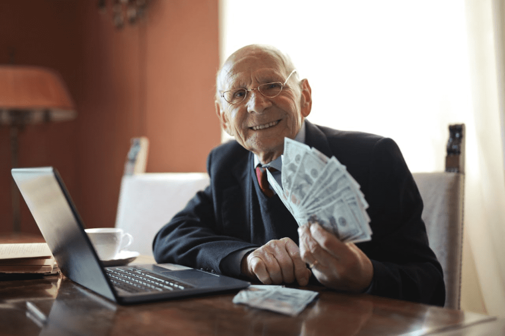 Happy senior businessman holding money in hand while working on laptop at table - Lead Grow Develop - Business, Technology, Personal Development Happy senior businessman holding money in hand while working on laptop at table