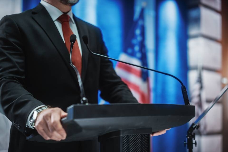 A man in a black suit and red tie stands at a miked podium, grabbing the edges with his hands. To his right is the US flag.