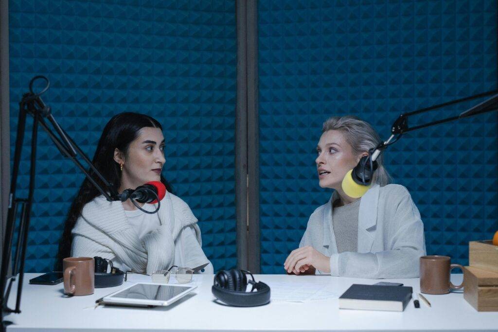 Two women sit at a podcast desk with microphones and headphones, chatting in a studio.