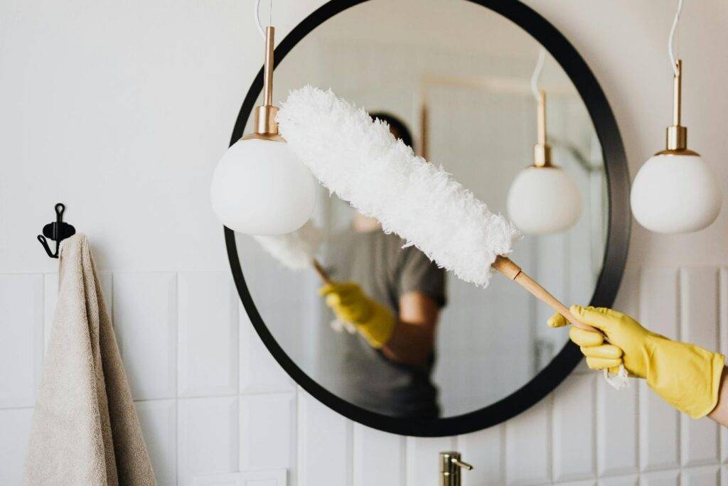 Person wearing yellow gloves dusting a round bathroom mirror with a white fluffy duster in view