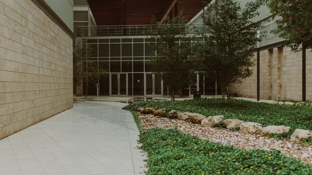 Modern building entrance with glass doors, beige stone walls, and a landscaped courtyard with trees and rocks.