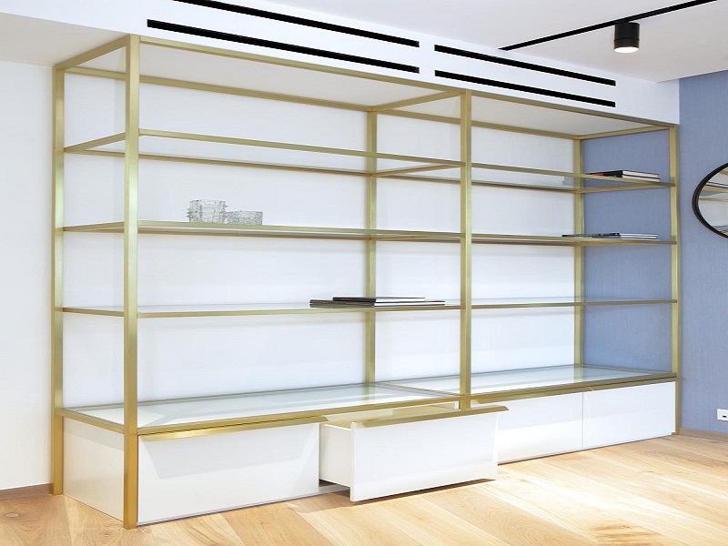 Gold metal frameless bookshelf with white drawers along the bottom in a bright living room, mostly empty shelves with a few books and decor pieces.